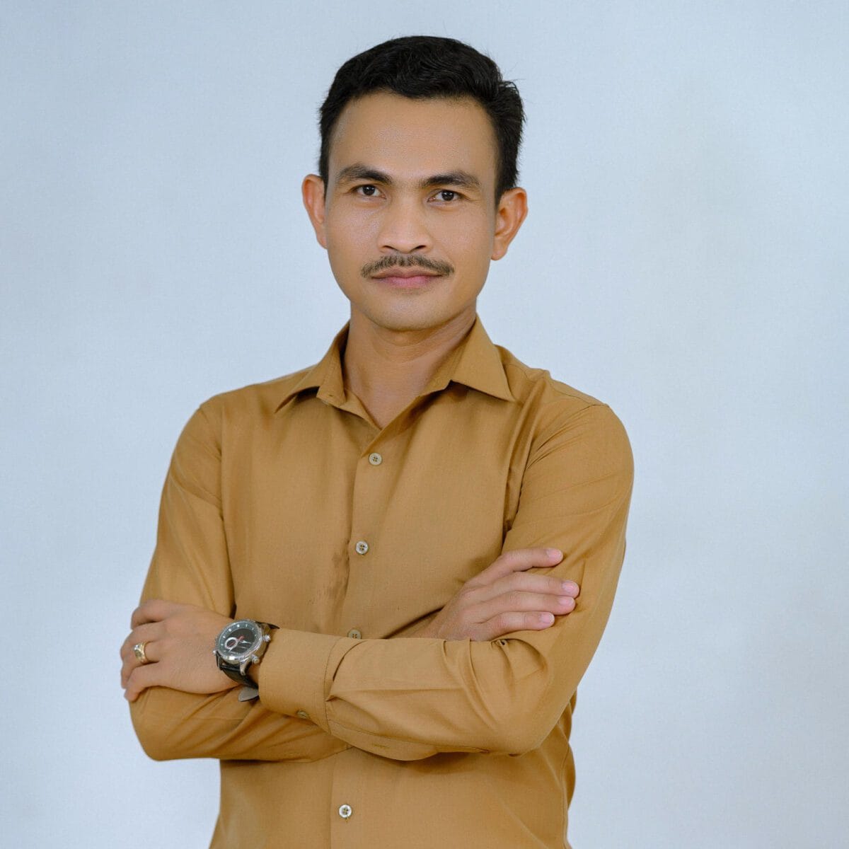 TDSO Confident male teacher with crossed arms, wearing a tan shirt, standing against a plain background.
