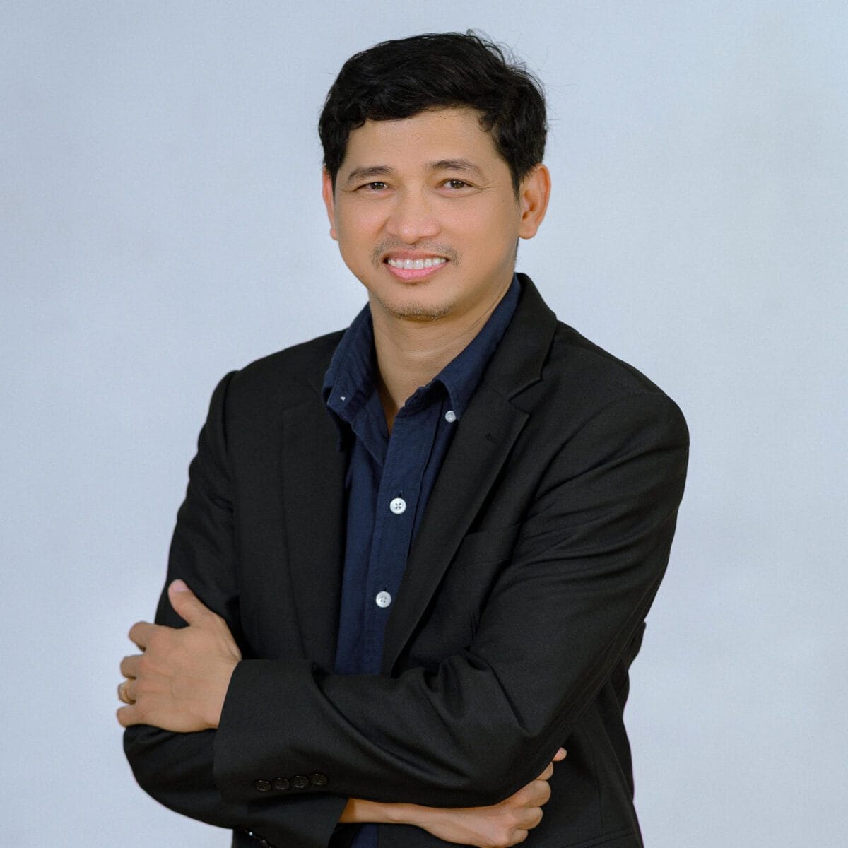 TDSO Professional educator smiling, wearing a black suit and blue shirt, standing confidently with arms c.